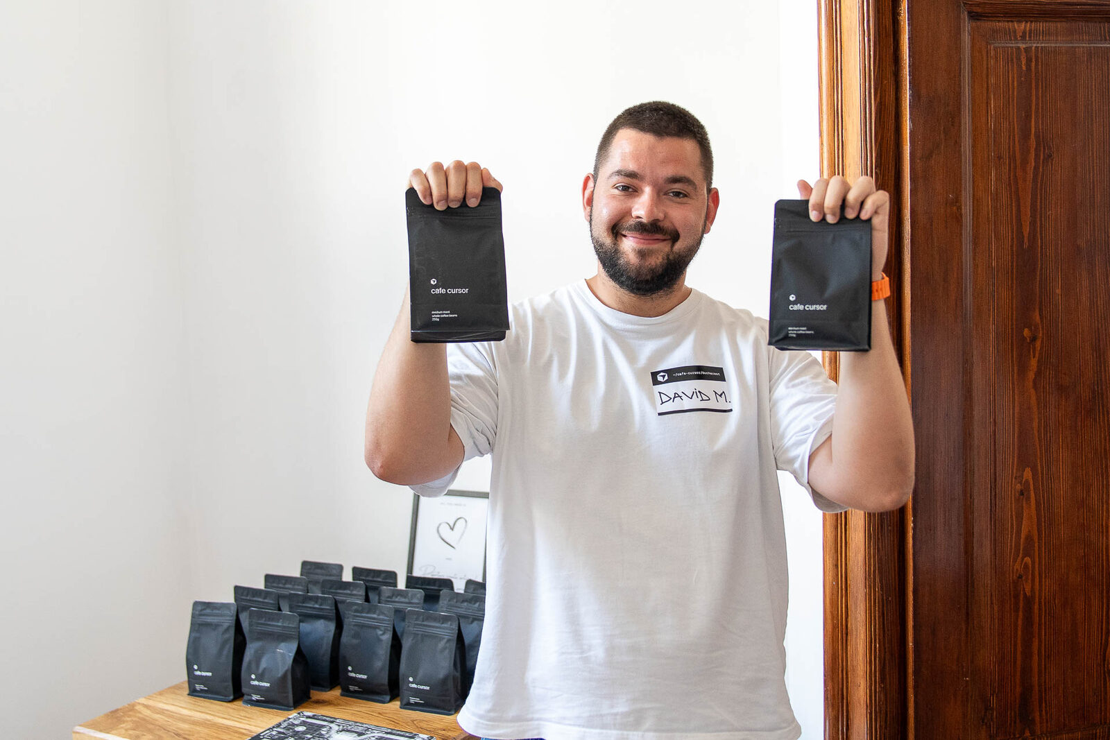 David Marin smiling, holding up two bags of Café Cursor branded coffee, a stack of more bags on the table in front of him.