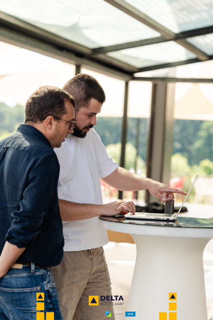Two founders working at a laptop on a standing table by a glass wall during Delta Bootcamp.