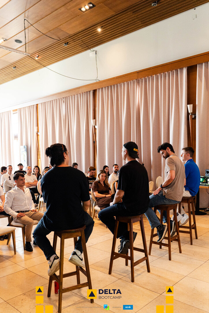 A bright room at Delta Bootcamp with founders seated on stools, listening to a mentor session.