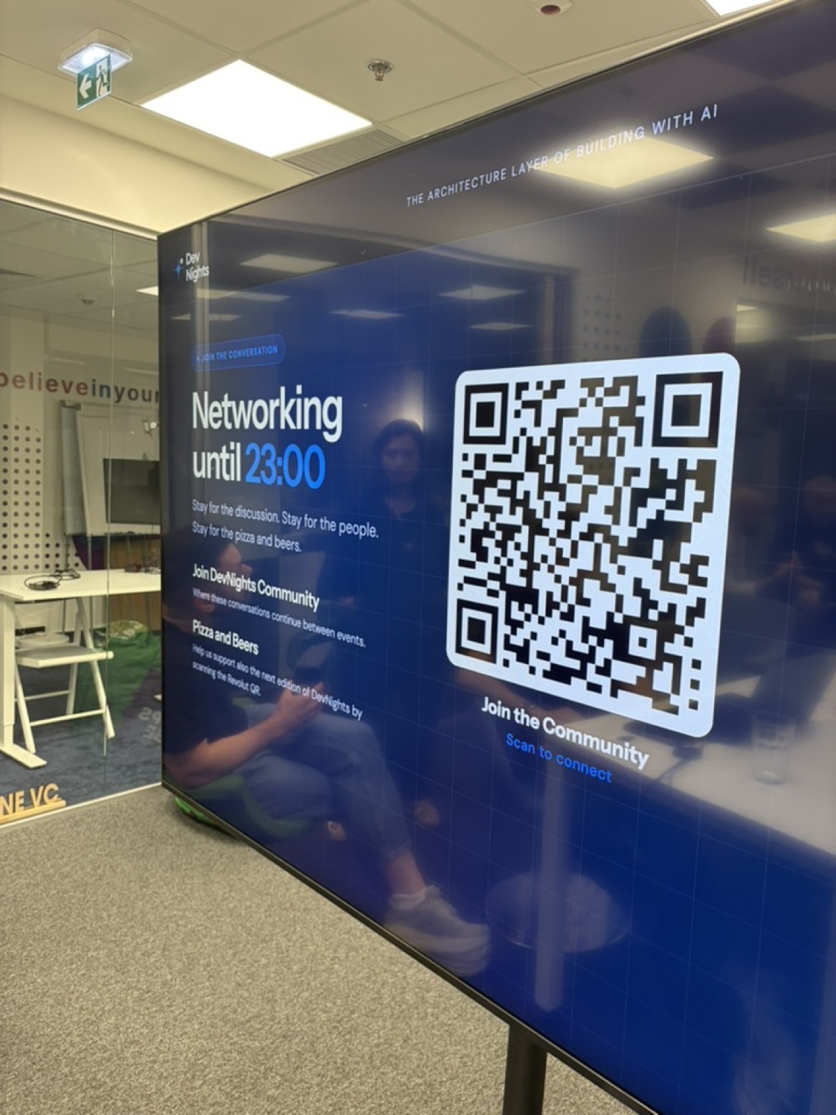 Un ecran la DevNights pe care scrie "Networking until 23:00. Stay for the discussion, stay for the people, stay for the pizza and beers.", lângă un cod QR pentru a intra în comunitate.