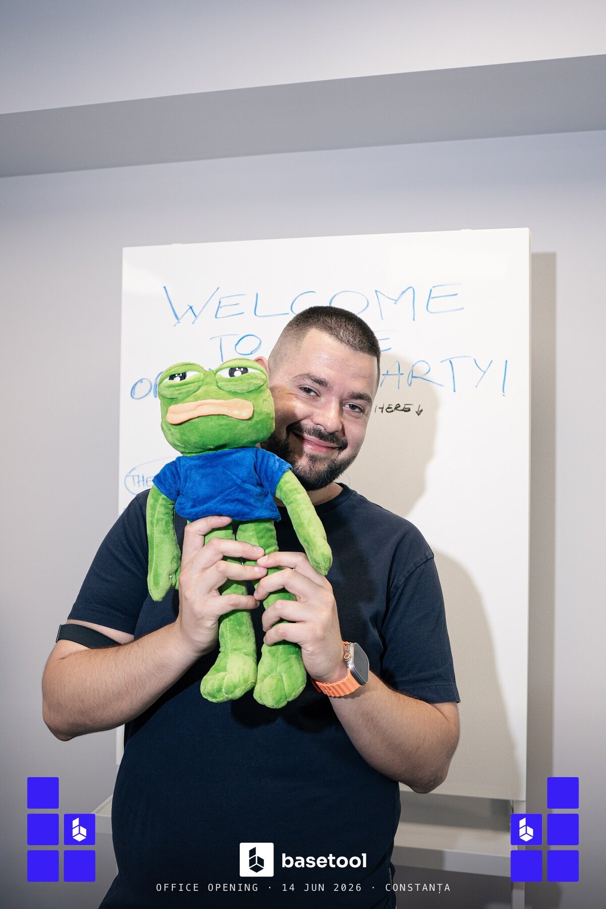 A teammate hugging Pepe, the studio's frog mascot, under the welcome-to-the-party whiteboard