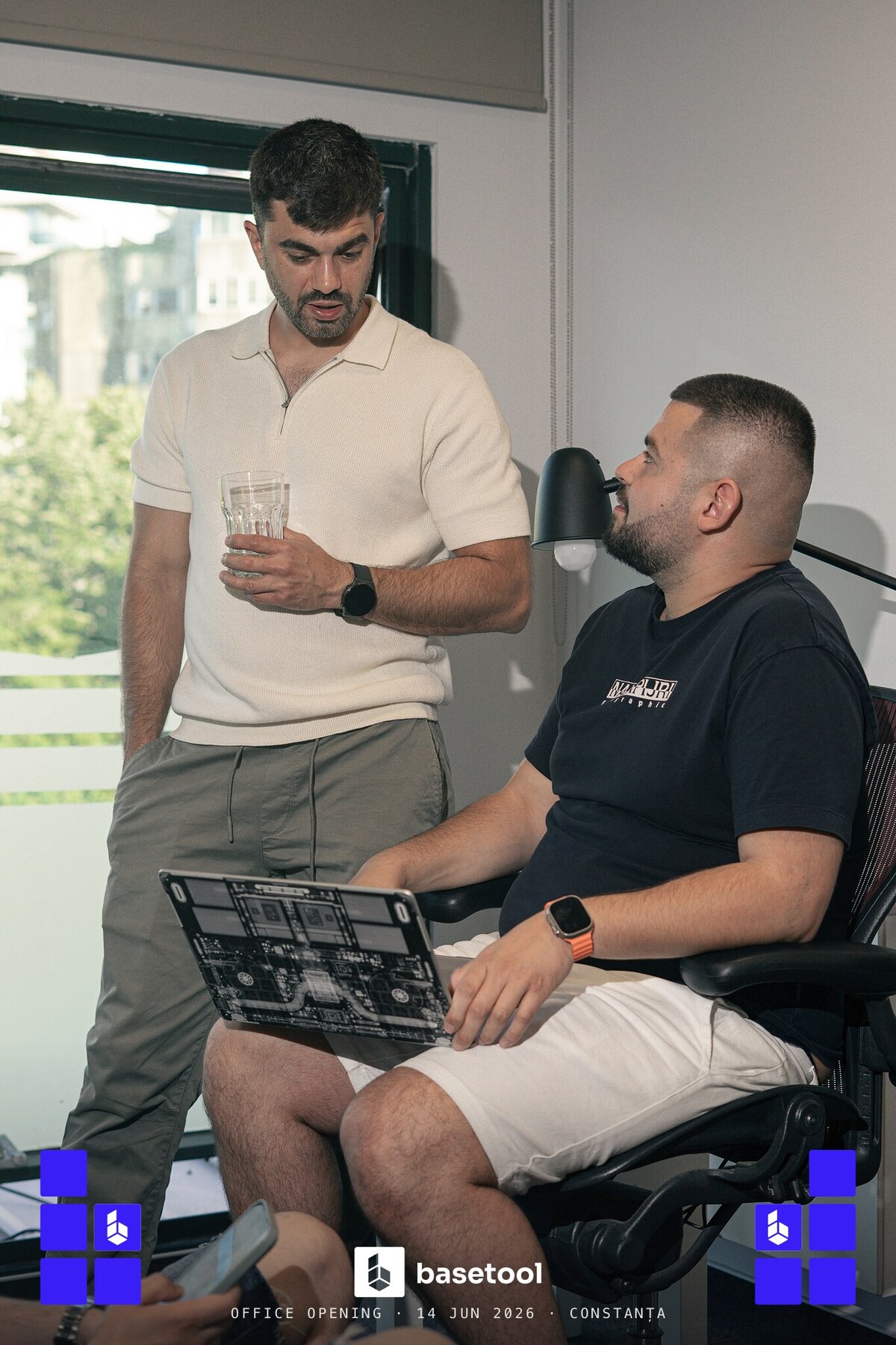 Two teammates leaning over a sticker-covered laptop while the party carries on
